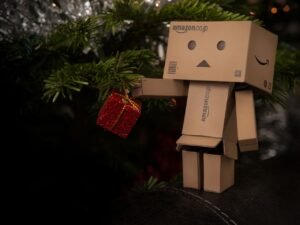 A cardboard figurine holds a small red gift near a decorated Christmas tree branch.